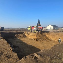 Firma Handlowo - Usługowa - Żółta koparka Volvo nabiera ziemię do czerwonej ciężarówki na placu budowy, gdzie wykopano długi rów pod fundamenty, w tle stoją domy i czerwona ciężarówka, a na skraju wykopu stoi geodeta z tyczką.