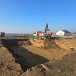 Żółto-czerwona koparka Volvo ładuje ziemię do czerwonej wywrotki na placu budowy wykopu pod fundamenty, w tle widoczny drugi samochód ciężarowy i budynki mieszkalne.