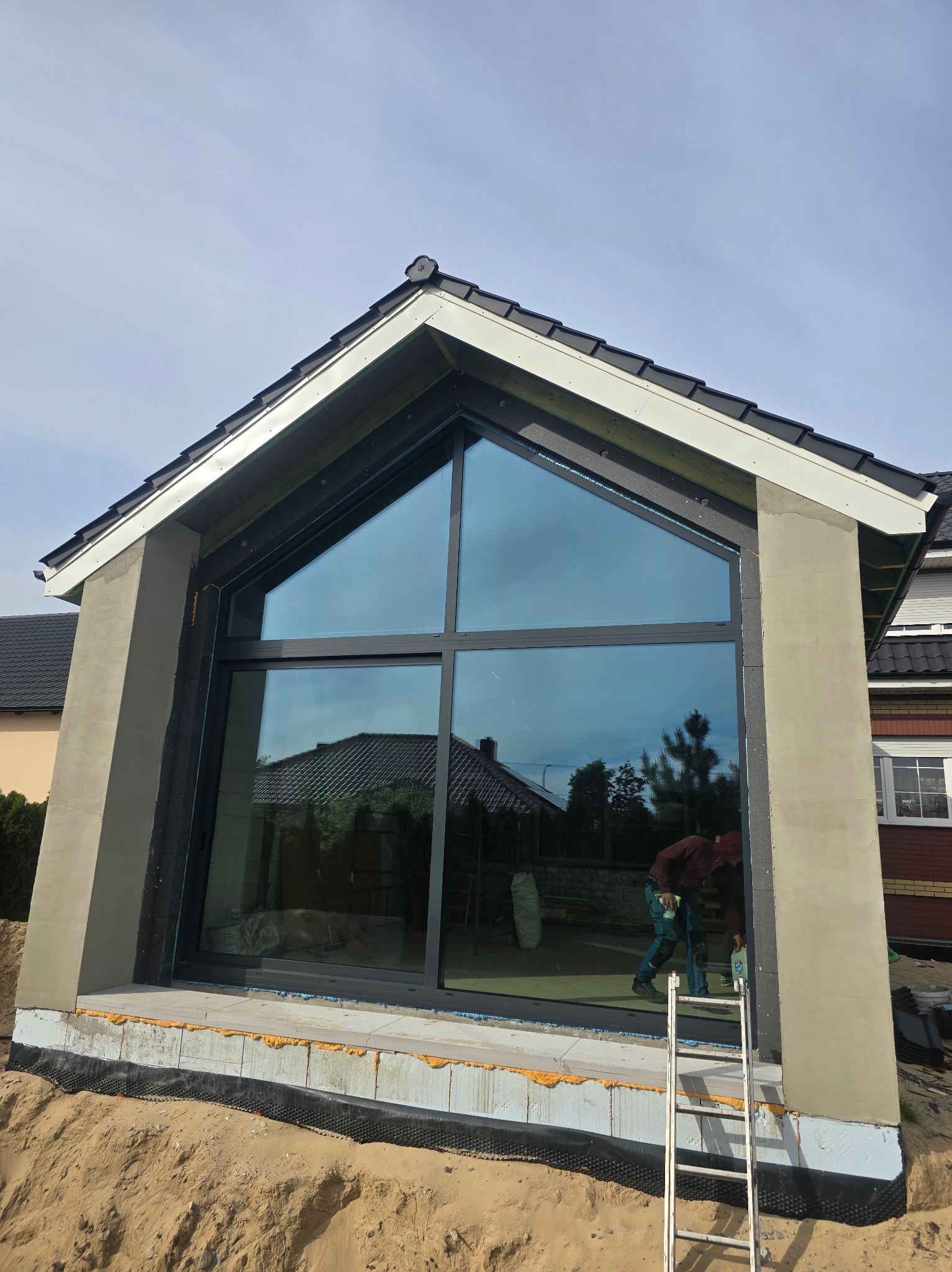 Montaż panoramicznego okna PCV w kształcie trapezu podczas wykańczania elewacji budynku, widoczny pracownik na drabinie, w tle odbicie sąsiedniego domu w szybie.