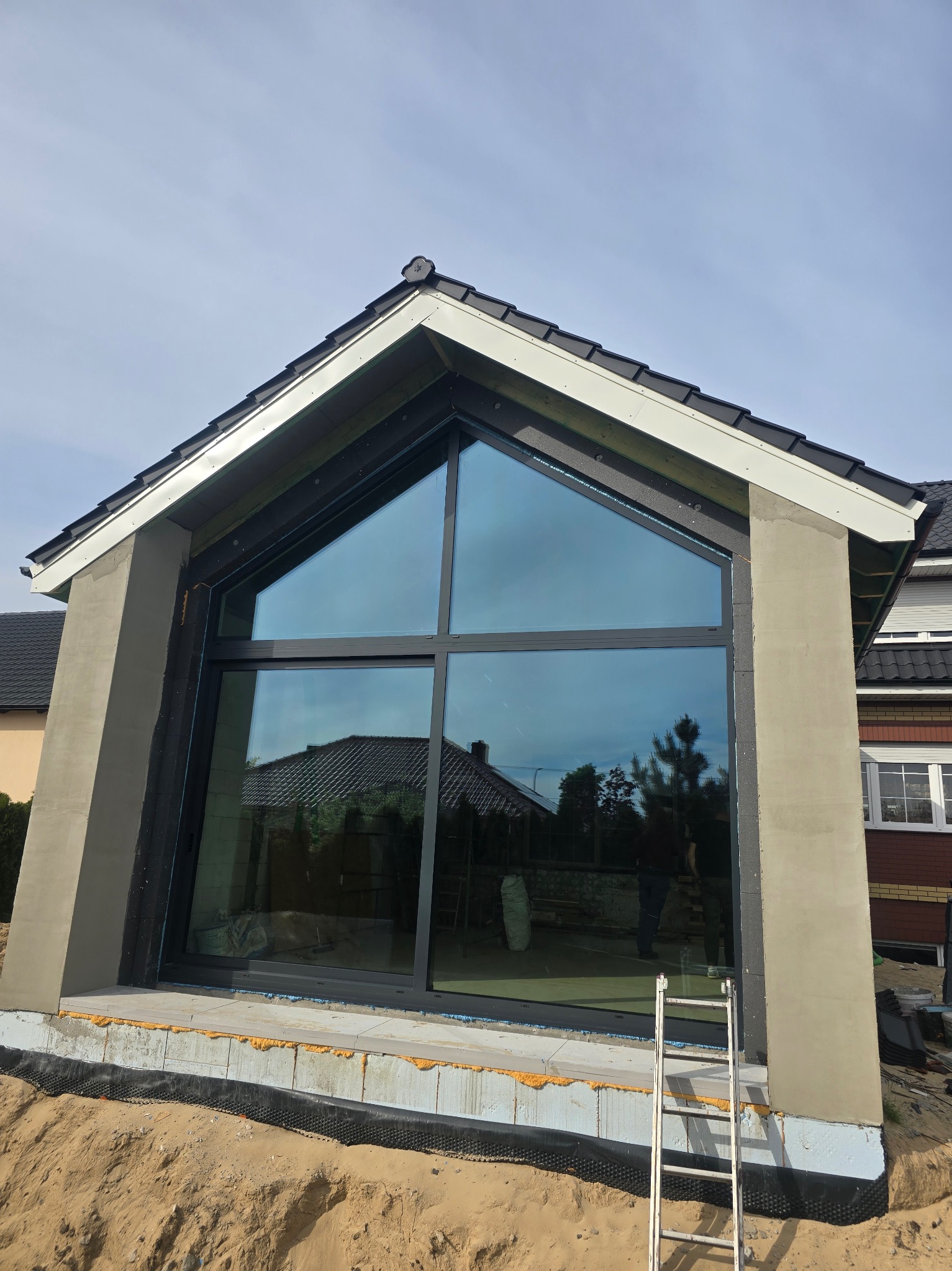 Nowoczesny dom w trakcie budowy z zamontowanym dużym trójkątnym oknem z czarną ramą, widoczna elewacja w surowym stanie i drabina oparta o ścianę.