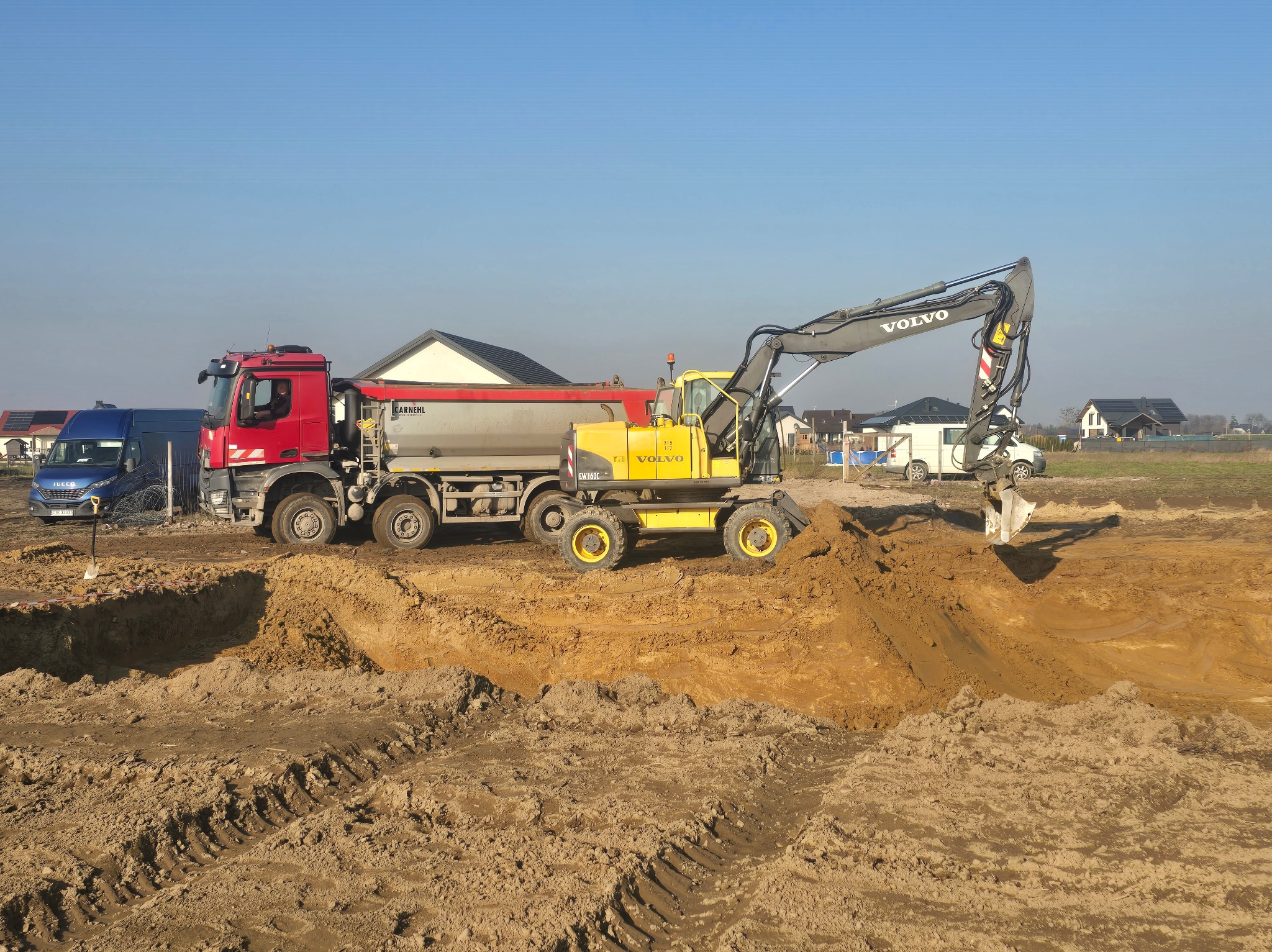 Żółta koparka Volvo i czerwona ciężarówka Carnehl pracują przy wykopie w Toruniu, na tle zabudowań mieszkalnych pod błękitnym niebem.