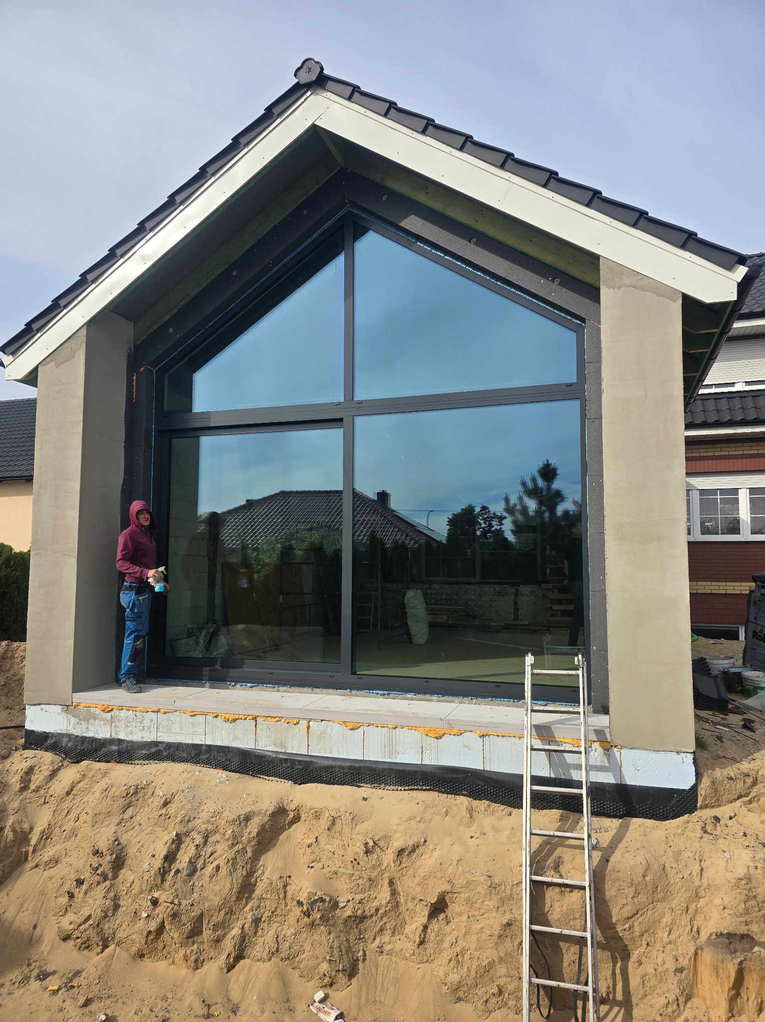 Montaż panoramicznego okna z czarną ramą w surowym stanie, widoczna drabina i pracownik z butelką płynu do czyszczenia szyb, odsłonięty fundament budynku w trakcie prac wykończeniowych.