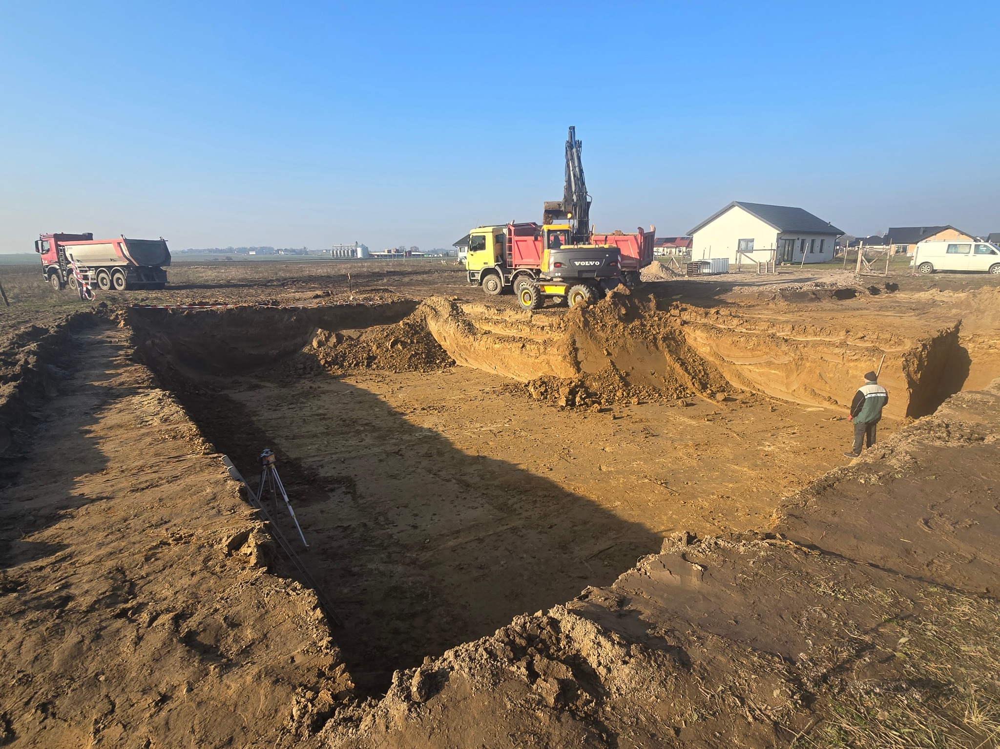 Głęboki wykop pod budowę, z widocznym sprzętem budowlanym: koparka Volvo na tle wywrotki i druga wywrotka w oddali, geodezyjny niwelator laserowy na statywie oraz pracownik w zielonej kurtce. W tle...