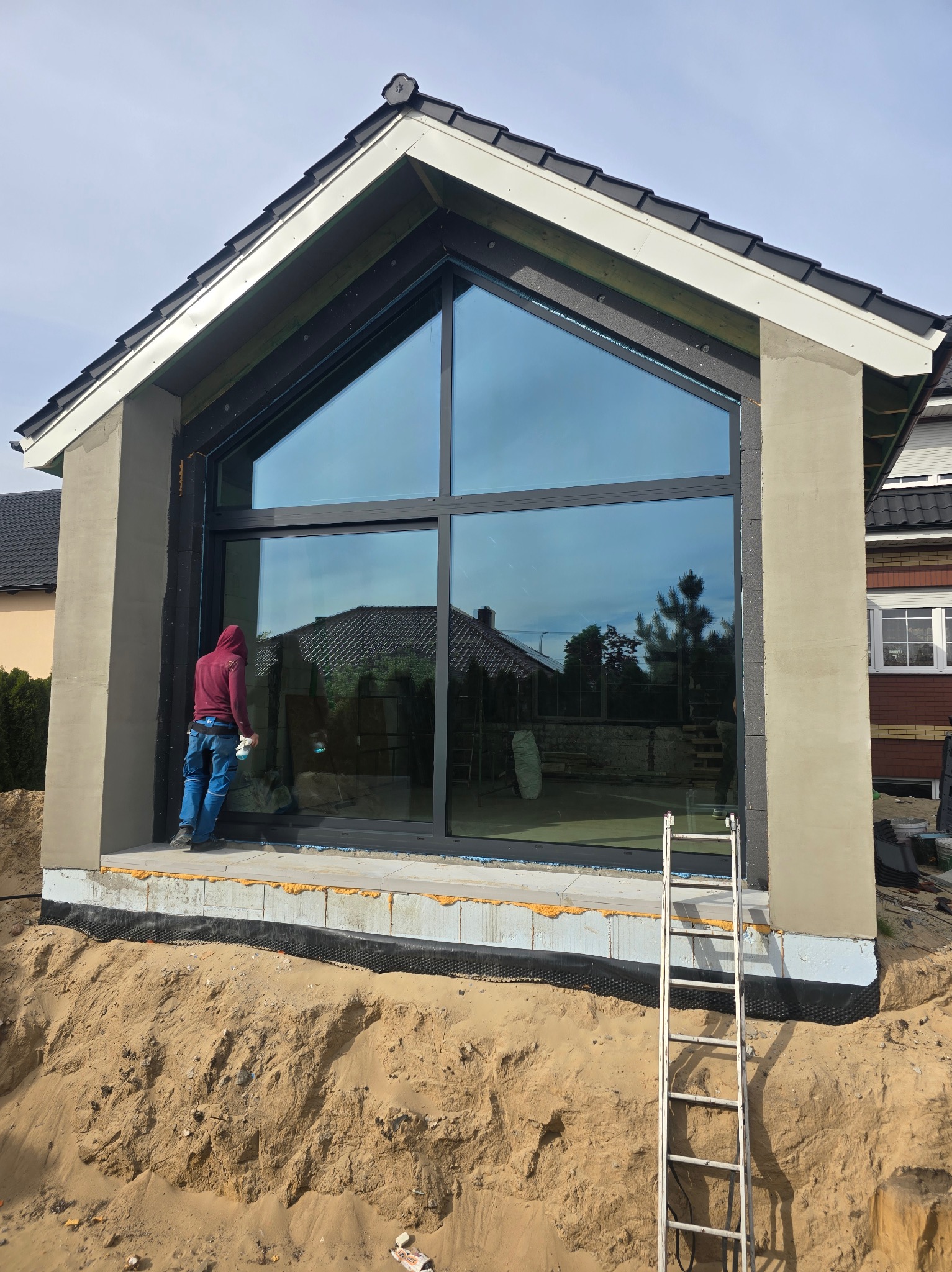 Montaż okna o dużej powierzchni w nowo budowanym domu, widoczny pracownik w czerwonej bluzie czyszczący szybę, odkryte fundamenty i drabina oparta o ścianę.