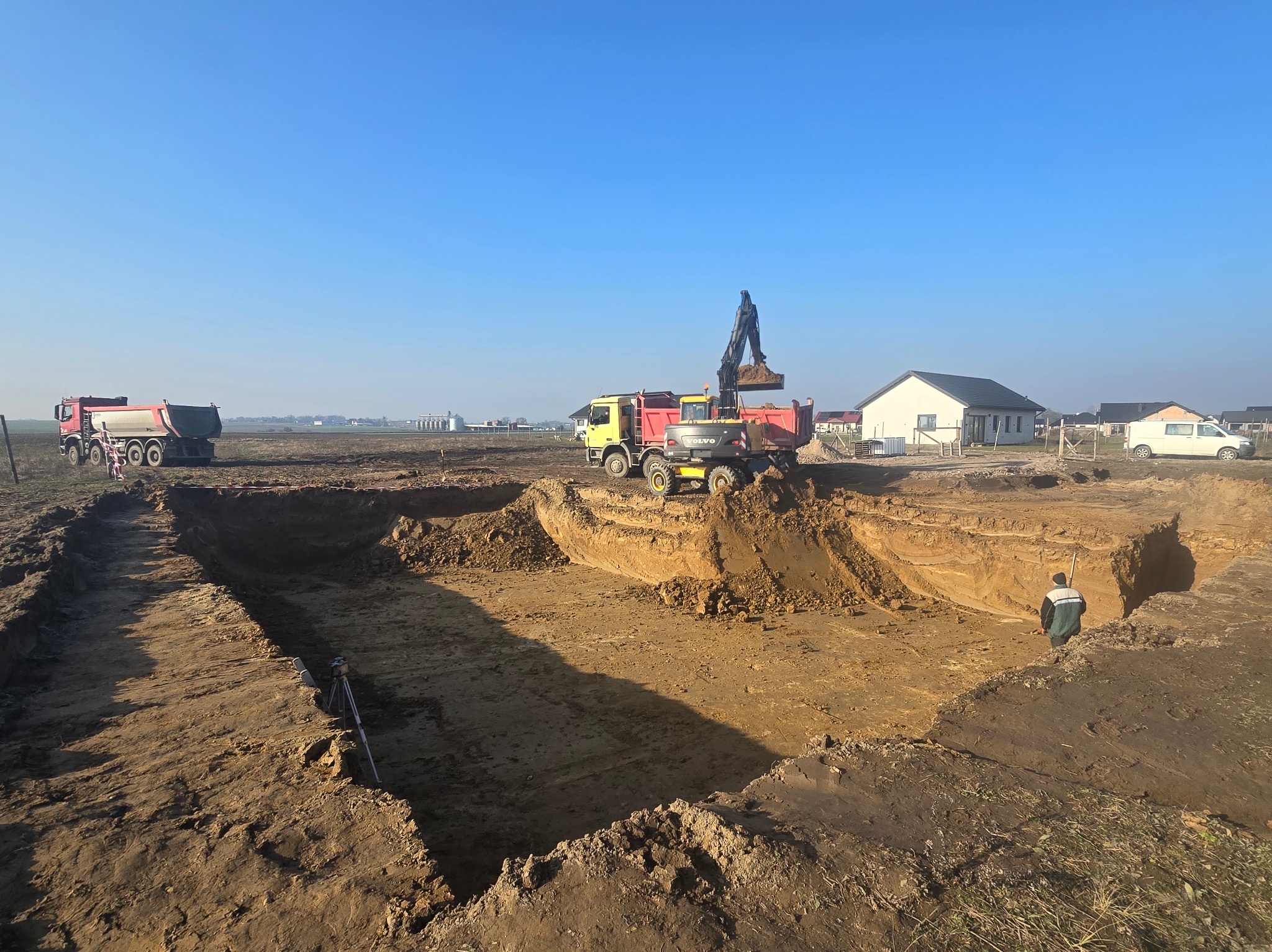 Żółto-czerwona koparka Volvo ładuje ziemię do czerwonej wywrotki na placu budowy wykopu pod fundamenty, w tle widoczny drugi samochód ciężarowy i budynki mieszkalne.