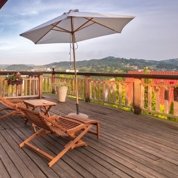 Piotr Zielonka - Agent Nieruchomości - Drewniany taras z widokiem na wzgórza, wyposażony w dwa leżaki, stolik, biały parasol przeciwsłoneczny i donice z kwiatami.