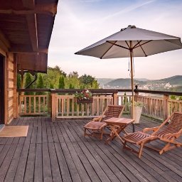 Piotr Zielonka - Agent Nieruchomości - Drewniany taras przy domu z bali, wyposażony w dwa leżaki, stolik i parasol, z widokiem na zielone wzgórza i zabudowania w oddali.