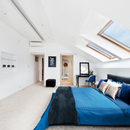 Piotr Zielonka - Agent Nieruchomości - Jasna sypialnia na poddaszu z dwoma oknami dachowymi, błękitna pościel na łóżku z czarnym zagłówkiem, stolik nocny z kwiatami i filiżankami, toaletka z lustrem i niebieskim krzesłem w tle.