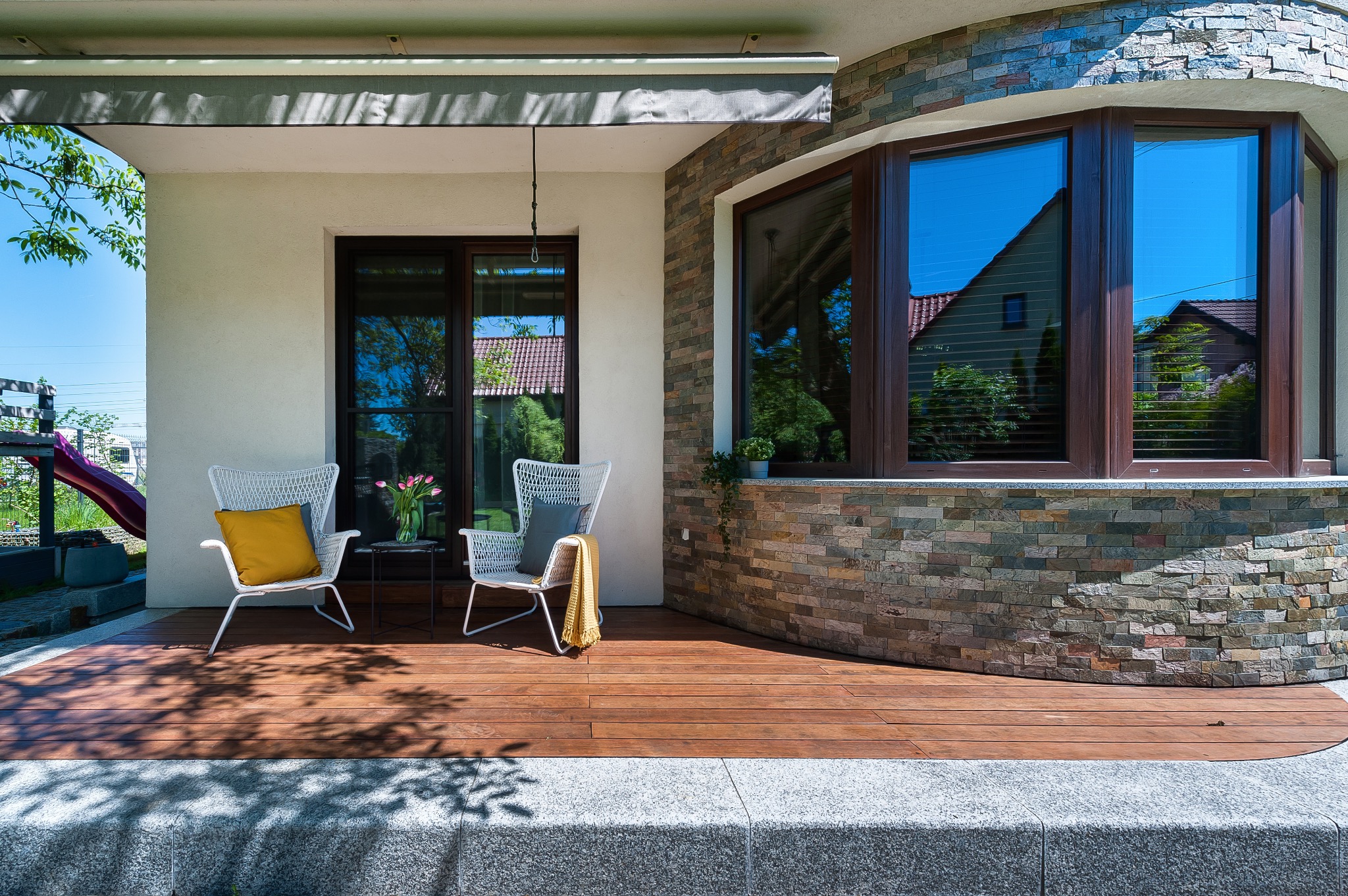 Zadaszony taras z drewnianą podłogą, wiklinowymi krzesłami z poduszkami, stolikiem z tulipanami i oknem z refleksami otoczenia.