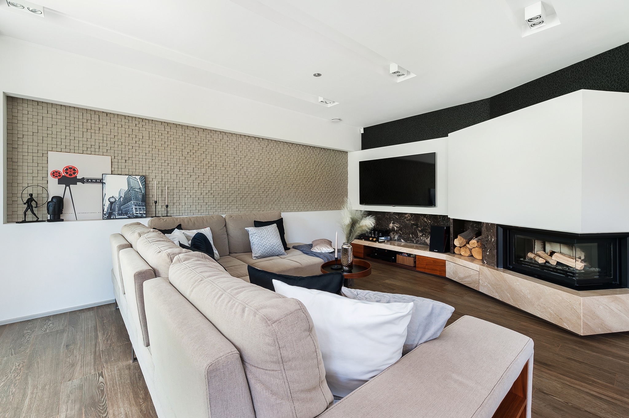 Nowoczesny salon z narożną sofą, kominkiem obudowanym marmurem i ścianą z dekoracyjnych paneli 3D, uzupełniony o telewizor i subtelne dekoracje.