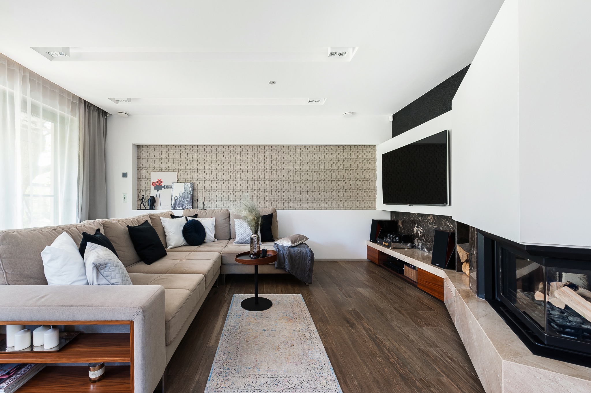 Jasny salon z narożną sofą, kominkiem z przeszklonym paleniskiem, telewizorem i ścianą z dekoracyjnej mozaiki. Na podłodze drewniany parkiet i dywanik.