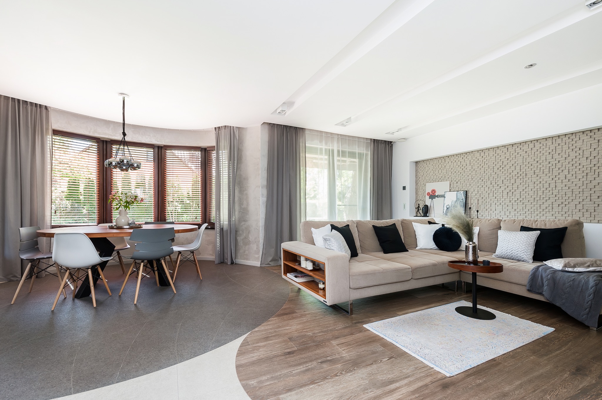 Jasne, przestronne wnętrze mieszkania z dużym oknem, okrągłym stołem jadalnianym i narożną sofą z dekoracyjnymi poduszkami, dywanem oraz mozaikową ścianą.