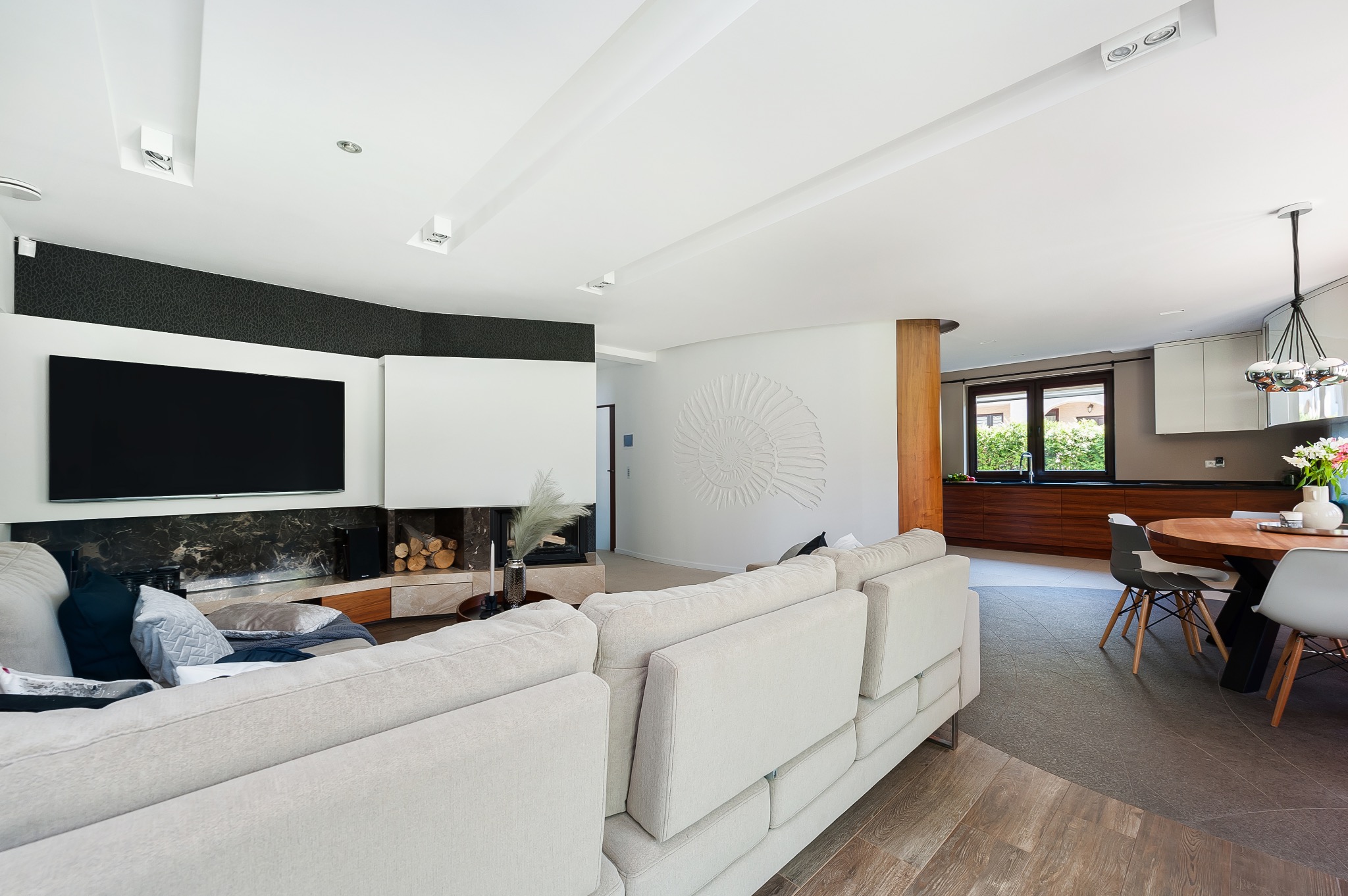 Jasny salon z beżową sofą, telewizorem nad kominkiem i jadalnią z okrągłym stołem w tle, widoczny element dekoracyjny w kształcie muszli na ścianie.