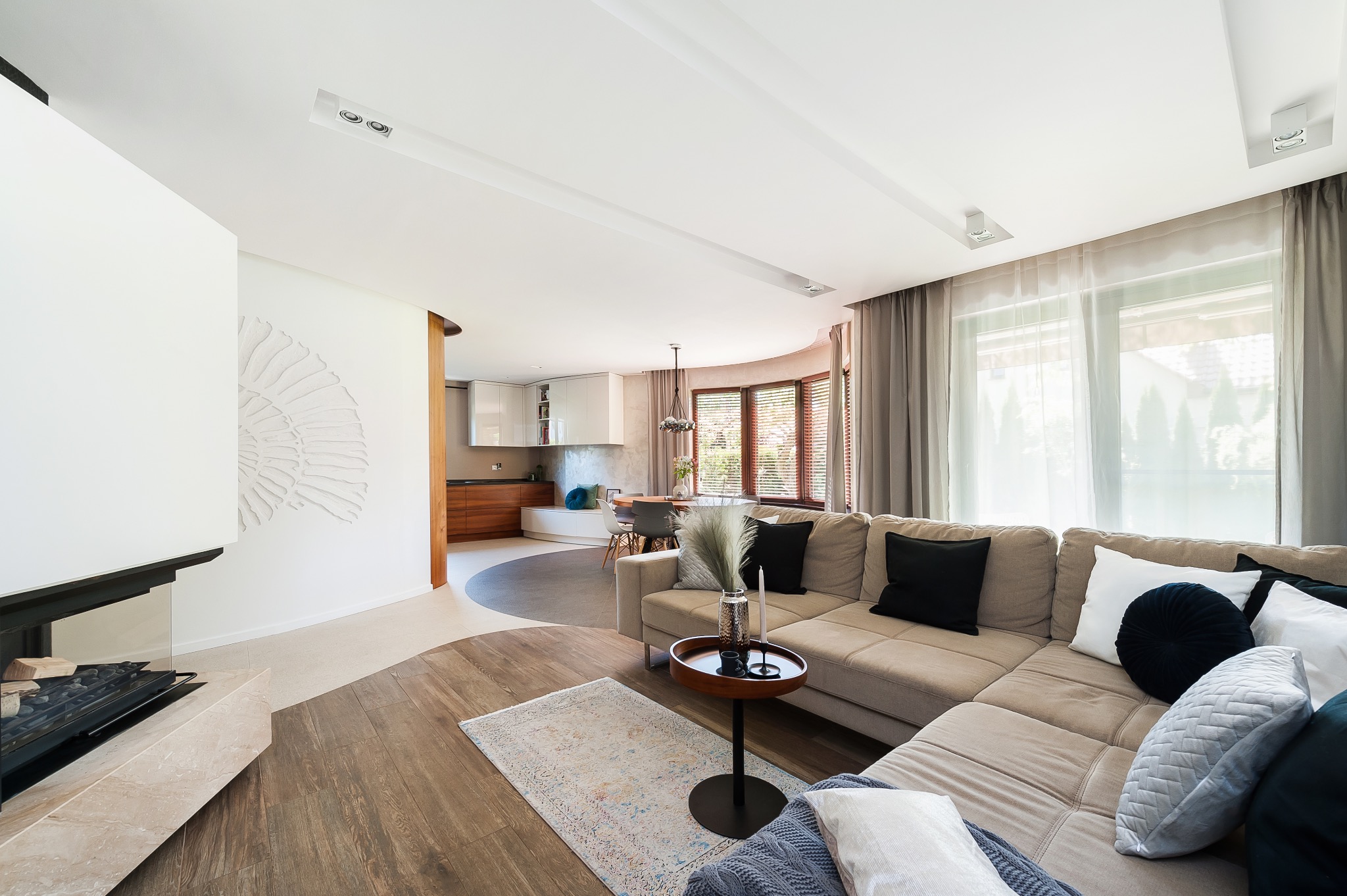 Jasne, przestronne wnętrze salonu z kominkiem, beżową sofą narożną z poduszkami, okrągłym stolikiem pomocniczym i widokiem na kuchnię z jadalnią w tle.