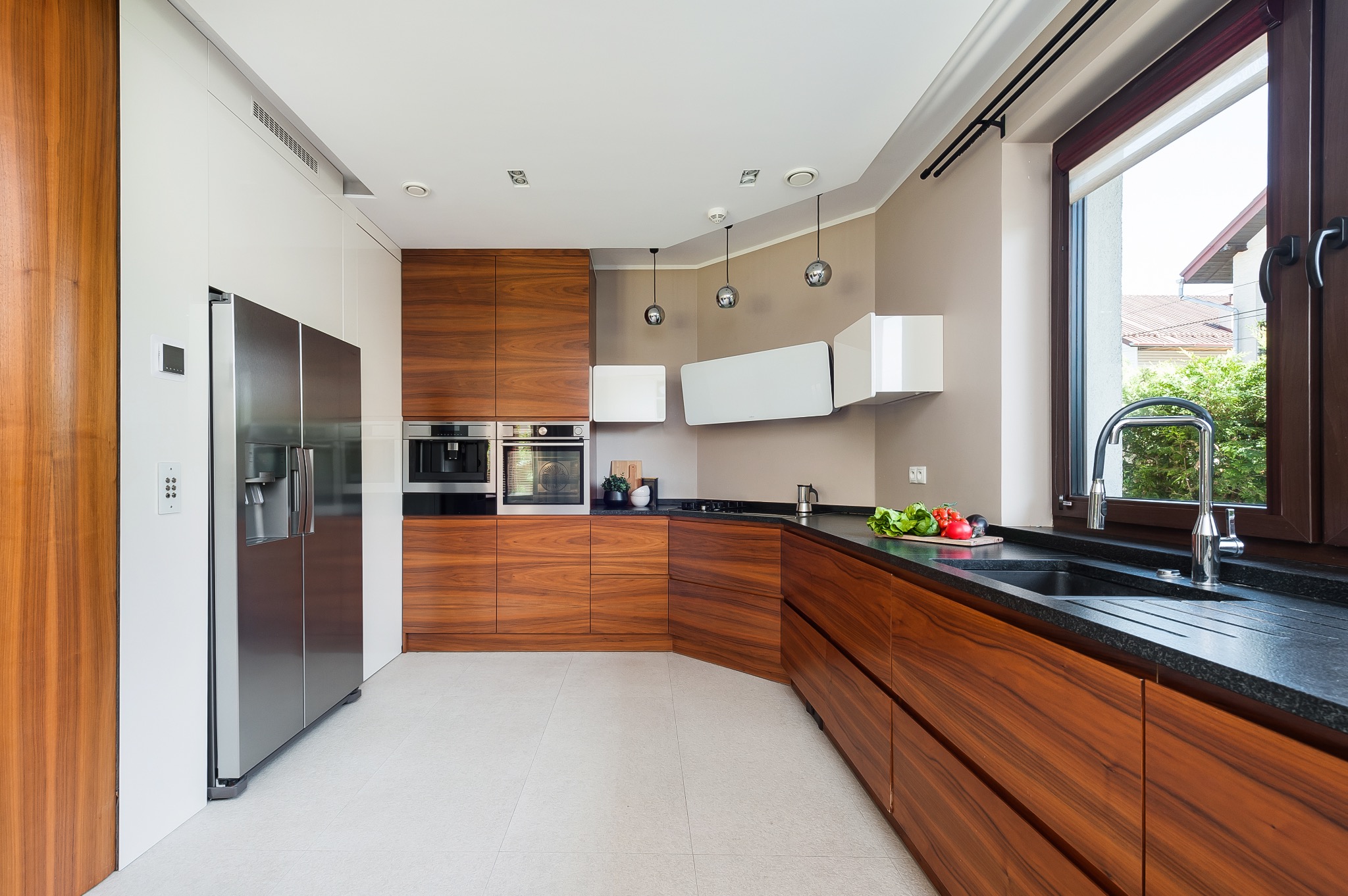 Nowoczesna kuchnia z drewnianymi frontami szafek, czarnym blatem i dużym oknem z widokiem na zieleń, oświetlona trzema wiszącymi lampami.