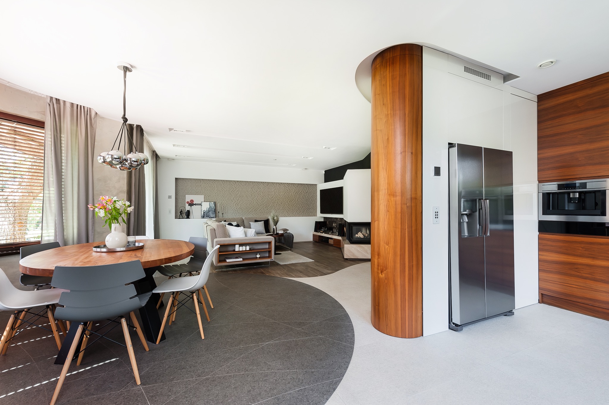 Wnętrze nowoczesnego apartamentu z otwartą przestrzenią, drewnianym filarem, okrągłym stołem jadalnym i designerską lampą wiszącą.