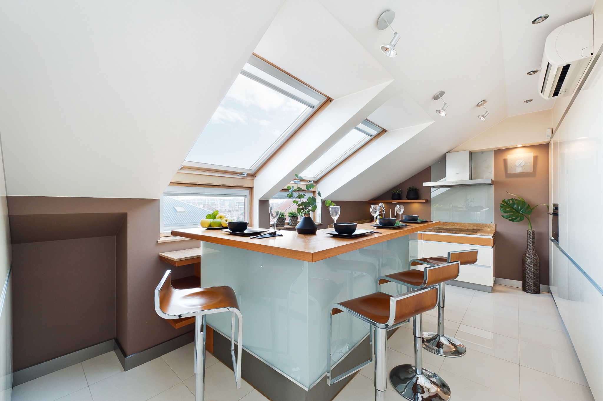 Nowoczesna kuchnia na poddaszu z wyspą, drewnianym blatem i czterema hokerami, oświetlona przez okna dachowe i punktowe lampy sufitowe.