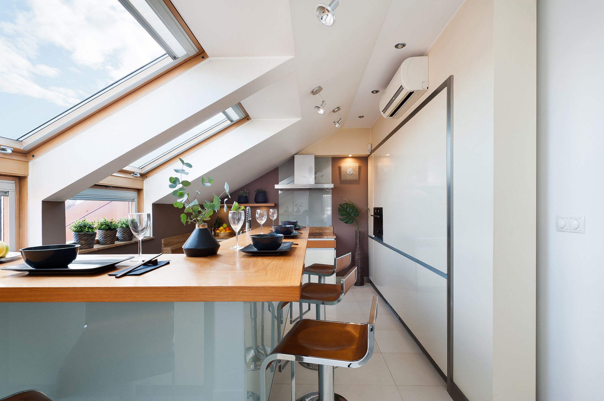 Nowoczesna kuchnia na poddaszu z drewnianym blatem, designerskimi krzesłami barowymi, przeszklonym dachem i minimalistycznymi szafkami, nakryta do posiłku dla kilku osób.