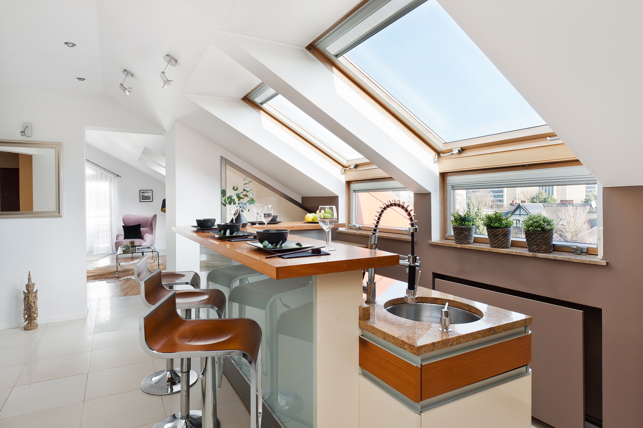 Jasna kuchnia na poddaszu z drewnianym blatem, hokerami, oknami dachowymi i widokiem na miasto z roślinami w doniczkach na parapecie.
