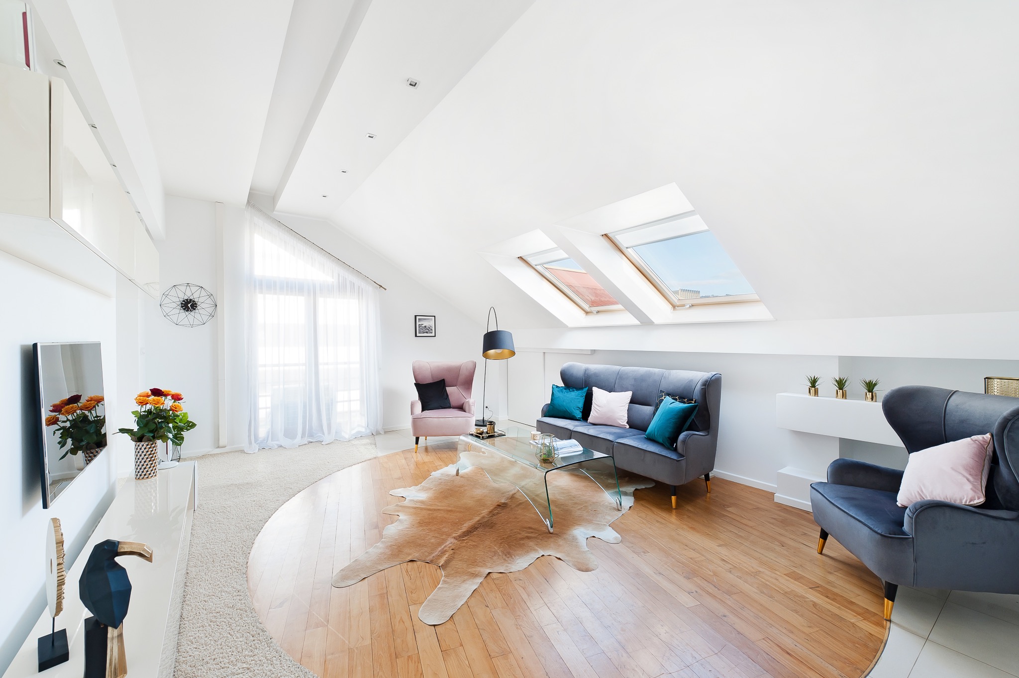 Jasne, przestronne poddasze z drewnianą podłogą, dywanem ze skóry zwierzęcej, szarym welurowym zestawem wypoczynkowym i oknami dachowymi w skosach.