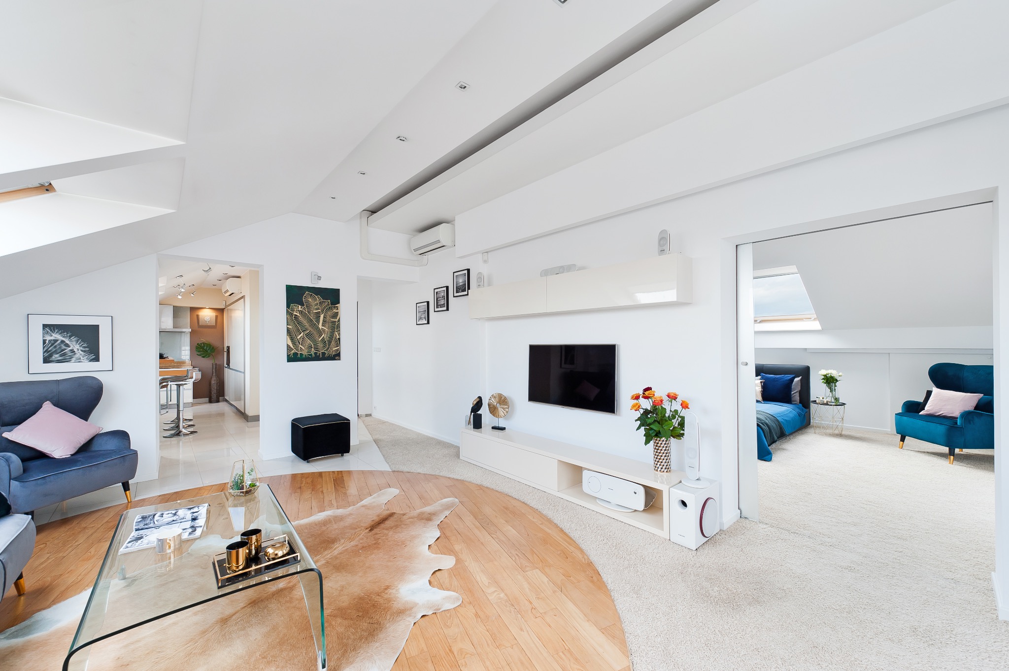 Jasne, przestronne wnętrze mieszkania z drewnianą podłogą i dywanem w kształcie skóry, szklanym stolikiem kawowym i telewizorem na ścianie, widok na sypialnię z niebieskimi akcentami i oknem dachowym.
