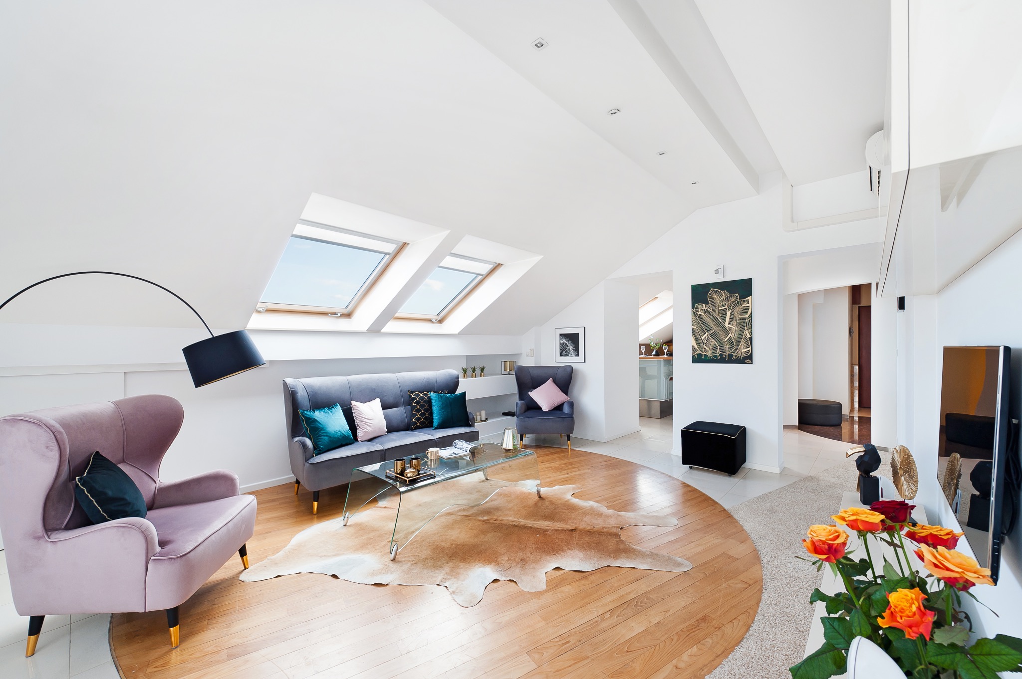 Jasne, przestronne poddasze w Krakowie z dwiema skośnymi oknami dachowymi, wyposażone w szary narożnik z kolorowymi poduszkami, szklany stolik kawowy i dekoracyjny dywan ze skóry. W tle widoczny...