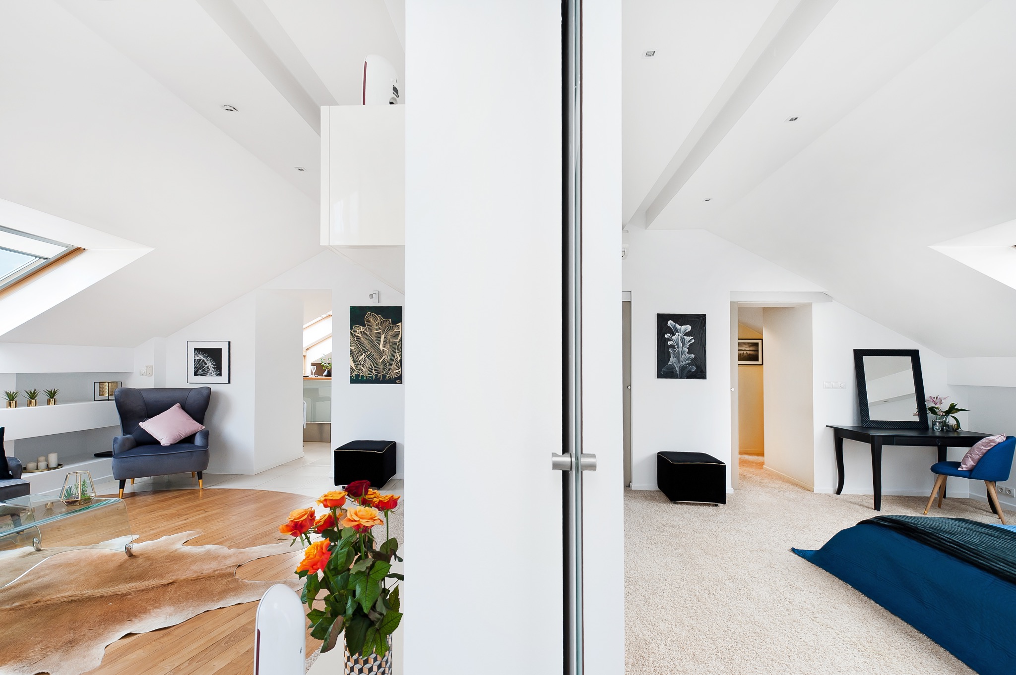 Dwupoziomowe, jasne mieszkanie na poddaszu z widokiem na salon z szarym fotelem i skórzanym dywanem oraz sypialnię z czarną toaletką i niebieskim łóżkiem, rozdzielone białymi drzwiami przesuwnymi.