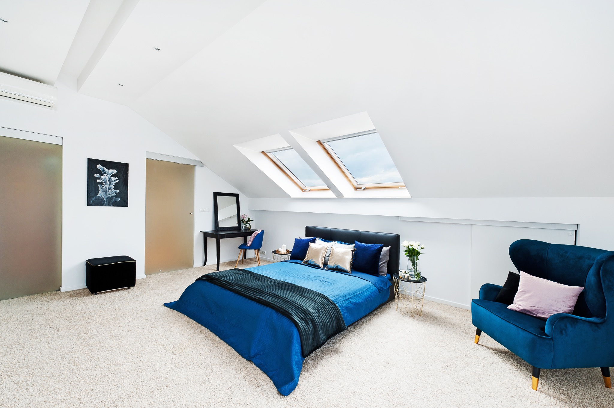 Jasna sypialnia na poddaszu z niebieskimi akcentami: łóżko z narzutą, fotel typu uszak i biurko pod oknem dachowym.