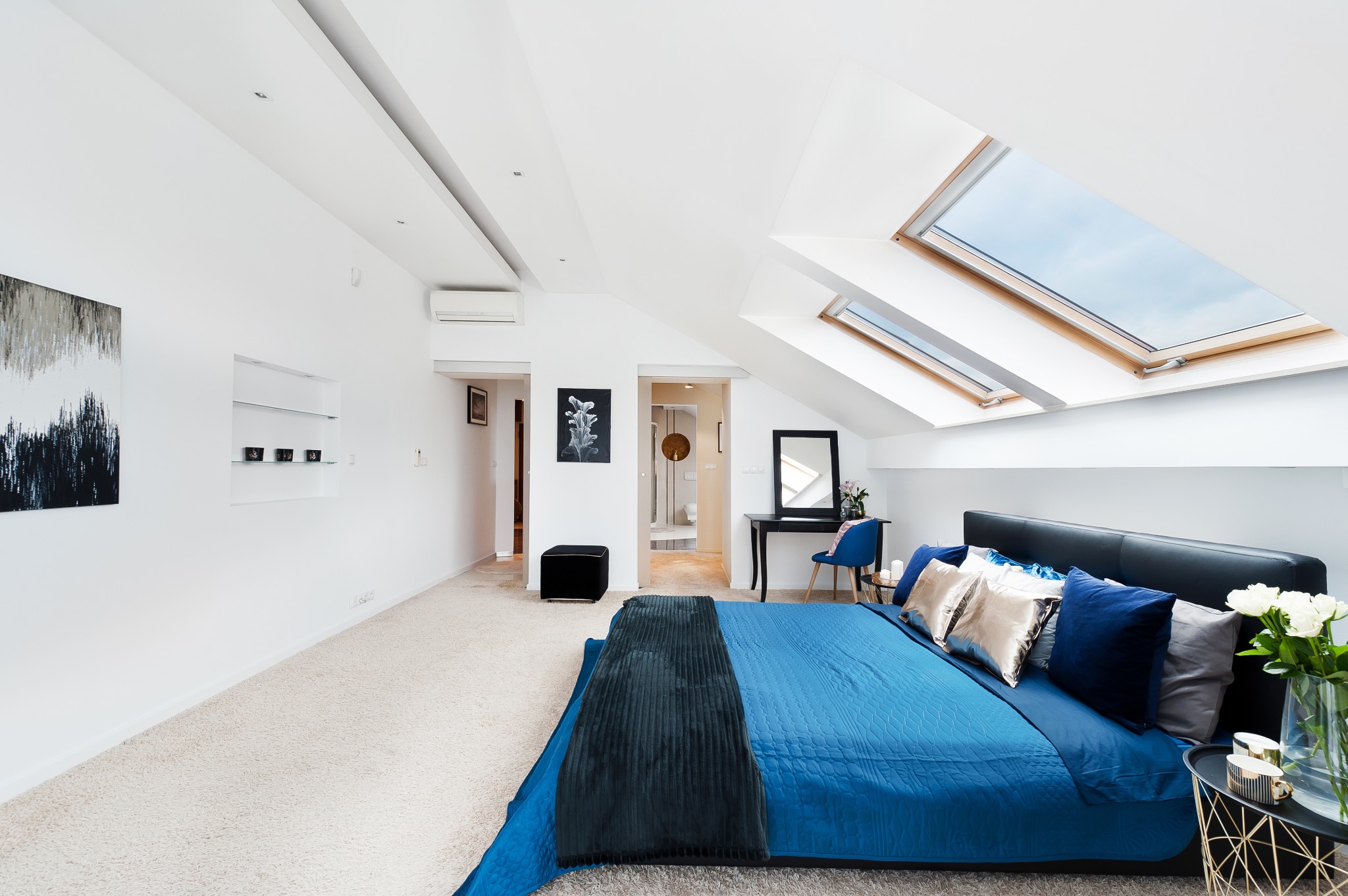 Jasna sypialnia na poddaszu z dwoma oknami dachowymi, błękitna pościel na łóżku z czarnym zagłówkiem, stolik nocny z kwiatami i filiżankami, toaletka z lustrem i niebieskim krzesłem w tle.