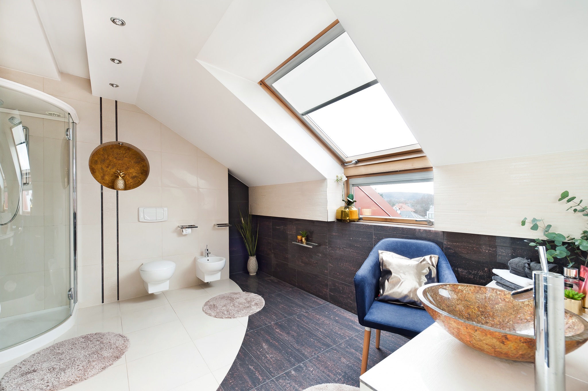 Jasna łazienka na poddaszu z kabiną prysznicową, bidetem, toaletą, umywalką w kształcie misy, krzesłem z poduszką, oknem dachowym i dekoracyjnymi elementami w kolorze złota.