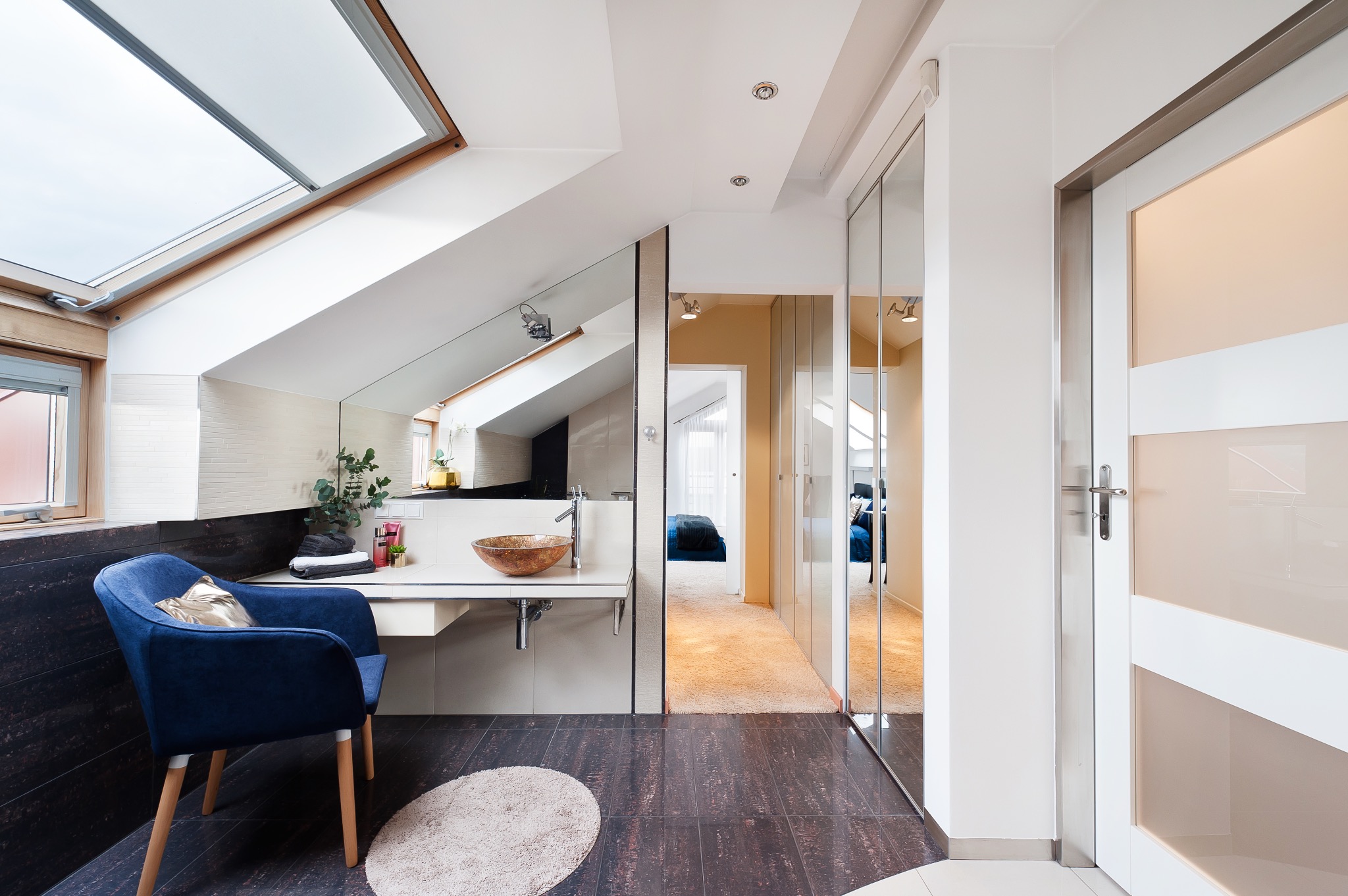 Jasna łazienka na poddaszu z drewnianą podłogą, umywalką w kształcie misy, lustrem i oknem dachowym, z widokiem na sypialnię.