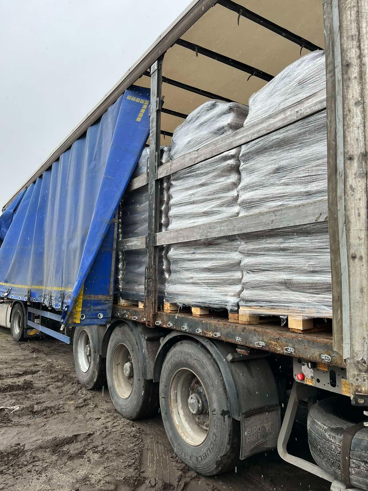 Ciężarówka z odkrytą plandeką, załadowana paletami z pelletem, widoczna w błotnistym terenie.