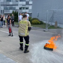 Strażak w umundurowaniu demonstruje gaszenie płonącej walizki na chodniku, grupa obserwatorów w tle, budynek biurowy.
