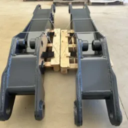 Dwie metalowe, pomalowane na szaro konstrukcje, prawdopodobnie ramiona koparki, leżą na drewnianej palecie, zabezpieczone białym sznurkiem.