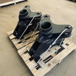 Dwie pomalowane na ciemno szaro metalowe konstrukcje, prawdopodobnie elementy maszyn, zabezpieczone białymi opaskami zaciskowymi i umieszczone na drewnianej palecie, na tle betonowej posadzki.