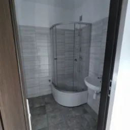 Wykończona łazienka z kabiną prysznicową narożną, umywalką i szarą glazurą imitującą kamień, widok zza uchylonych drzwi.