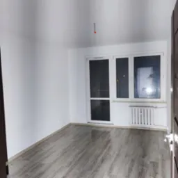 Wnętrze pustego, pomalowanego na biało pokoju w Siemirowicach z panelami podłogowymi, widoczne wystające kable ze ściany i grzejnik pod oknem.