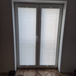 Grzegorz Borek - Okno strychowe z zamontowanymi białymi roletami plisowanymi. Na podłodze widoczne narzędzia i kable, sugerujące niedawny montaż.