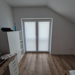 Grzegorz Borek - Jasne, podwójne okno strychowe z zamontowanymi białymi roletami plisowanymi. Na podłodze panele imitujące drewno. Biała komoda z akcesoriami do montażu.