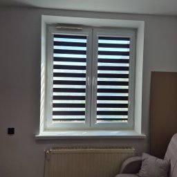 Grzegorz Borek - Okno z roletami dzień noc w białej ramie, widok z wnętrza. Na parapecie cień, pod oknem grzejnik, obok fragment sofy. Minimalistyczny wystrój.