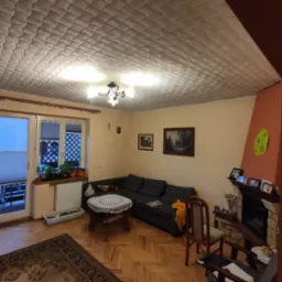 Salon z szarą sofą, kominkiem z cegły, oknem z roletami plisowanymi i kasetonowym sufitem podwieszanym.