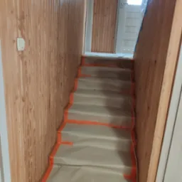 Schody zabezpieczone folią malarską i taśmą przed remontem, ściany wyłożone boazerią, widoczny fragment drzwi z przeszkleniami.