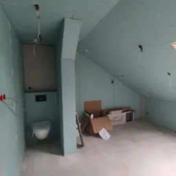 Adaptacja poddasza: ściany i sufit obłożone płytami gipsowo-kartonowymi w kolorze zielonym, widoczne instalacje elektryczne, zabudowa toalety w trakcie montażu, kartony z wyposażeniem sanitarnym...