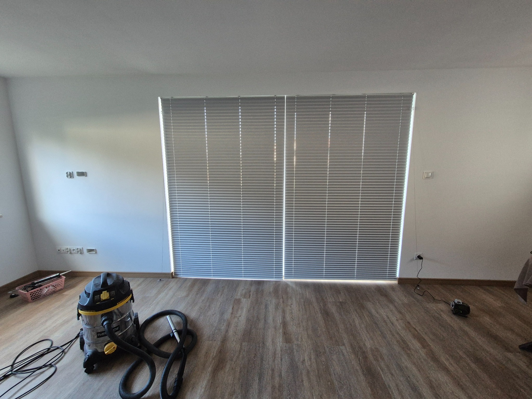 Jasne, aluminiowe żaluzje okienne zamontowane w oknie. Na podłodze widoczny odkurzacz przemysłowy i akcesoria, sugerujące prace porządkowe po instalacji.