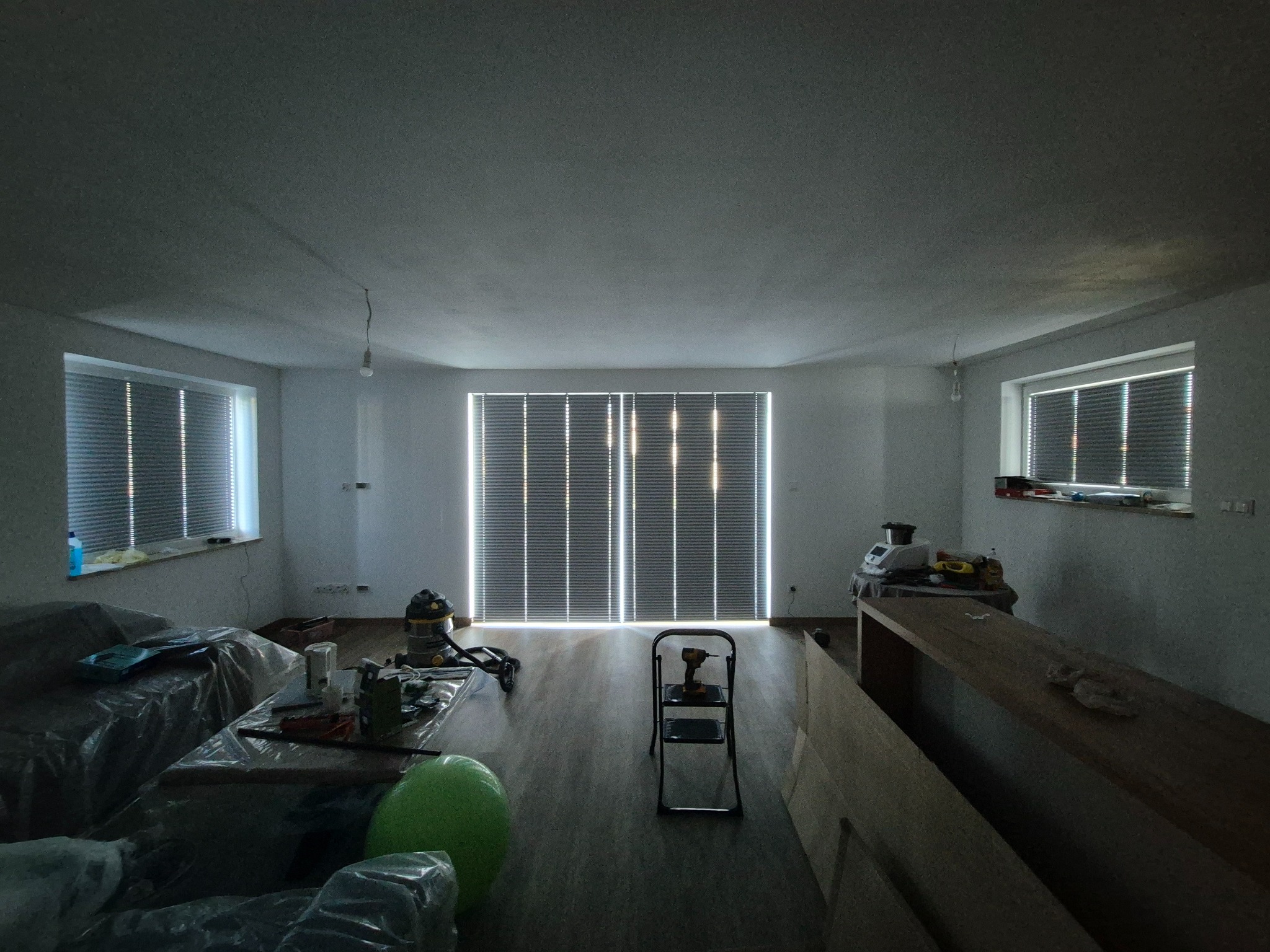 Pokój w trakcie remontu: widoczne okna z żaluzjami, drabina z wiertarką, zabezpieczone meble folią, podłoga w trakcie układania. Oświetlenie naturalne.