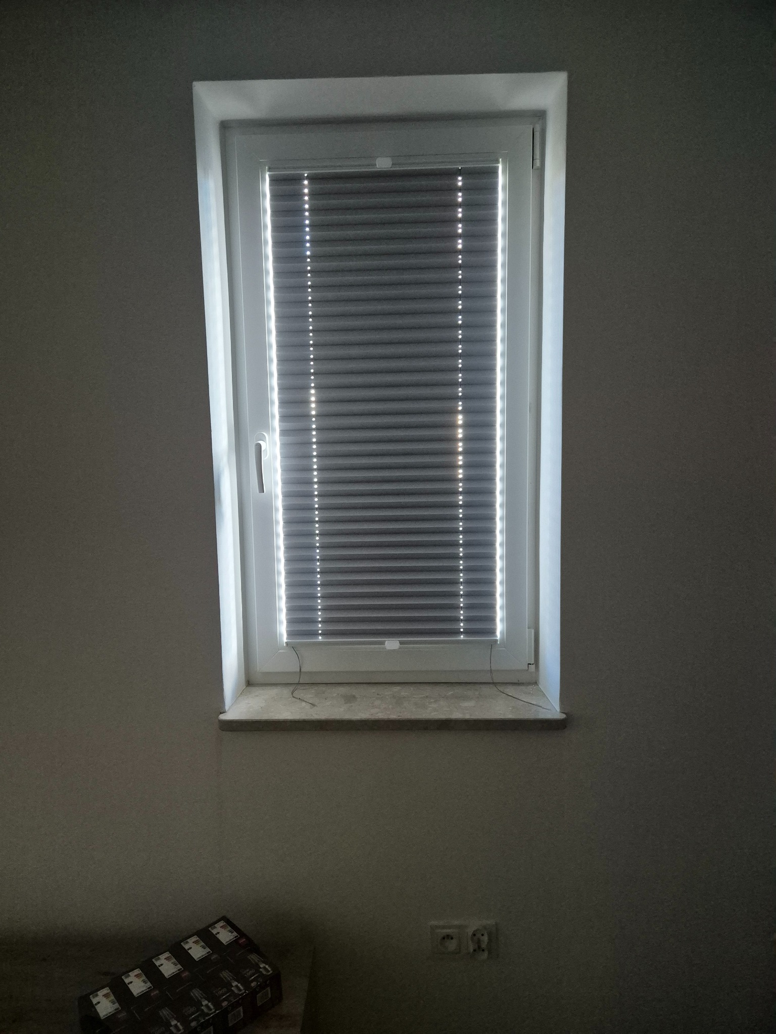 Zamknięte rolety dzień noc w białej ramie okiennej, widoczne na tle szarej ściany. Na parapecie widoczne przewody. W dolnej części kadru kartonowe opakowania.