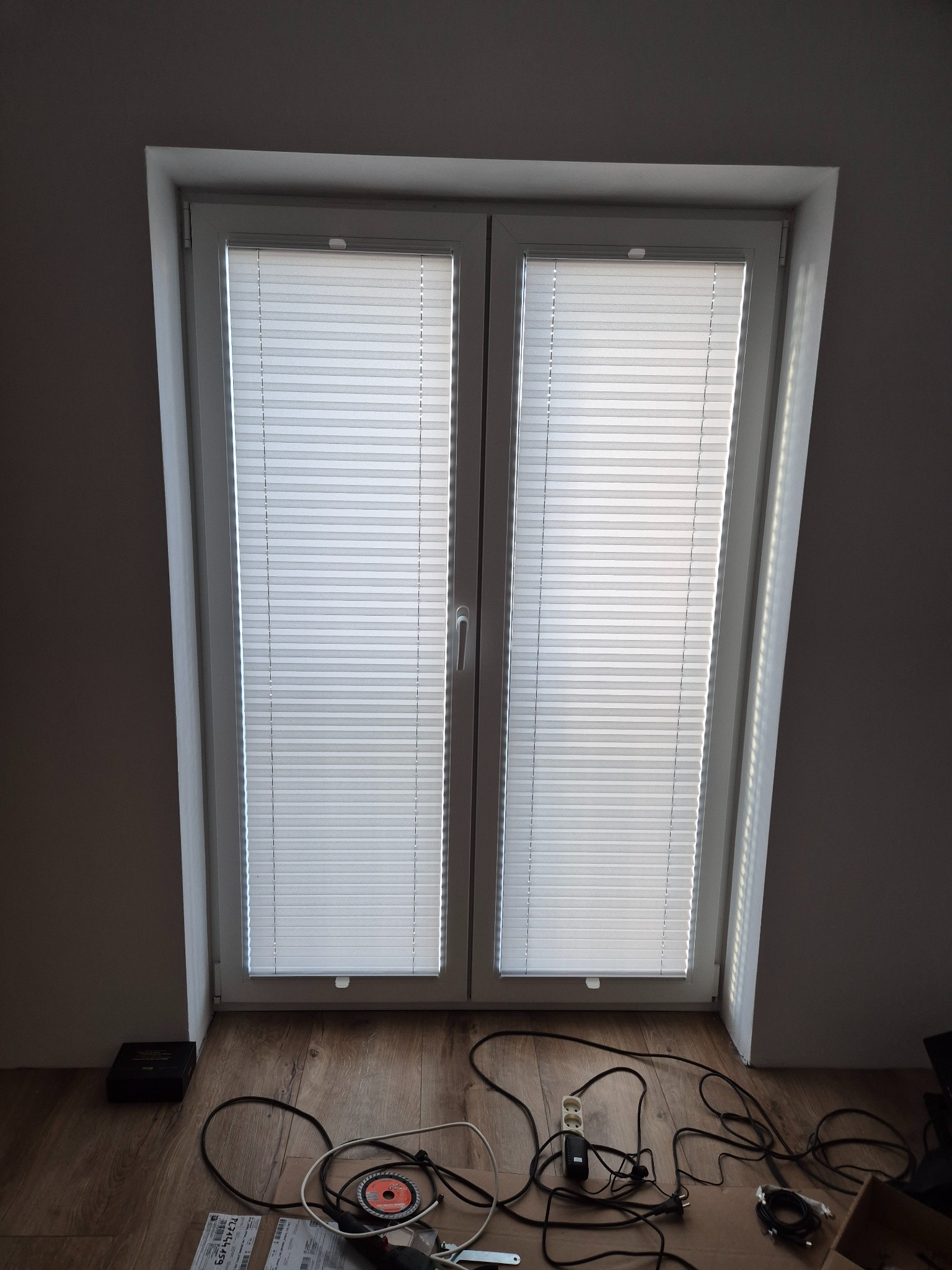 Okno strychowe z zamontowanymi białymi roletami plisowanymi. Na podłodze widoczne narzędzia i kable, sugerujące niedawny montaż.