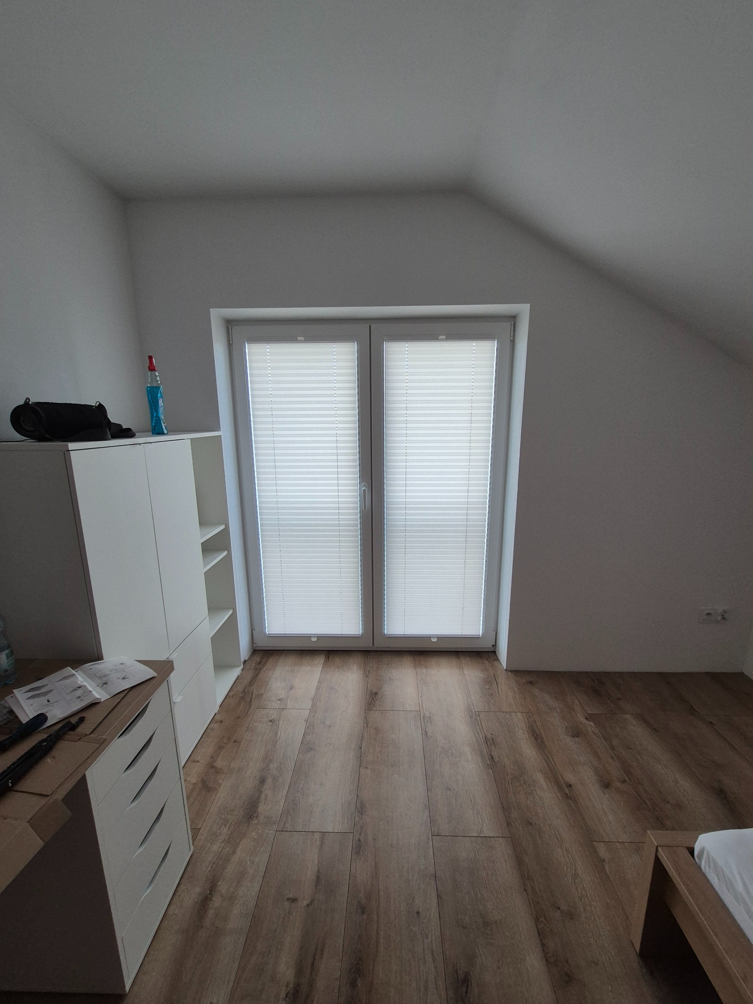 Jasne, podwójne okno strychowe z zamontowanymi białymi roletami plisowanymi. Na podłodze panele imitujące drewno. Biała komoda z akcesoriami do montażu.
