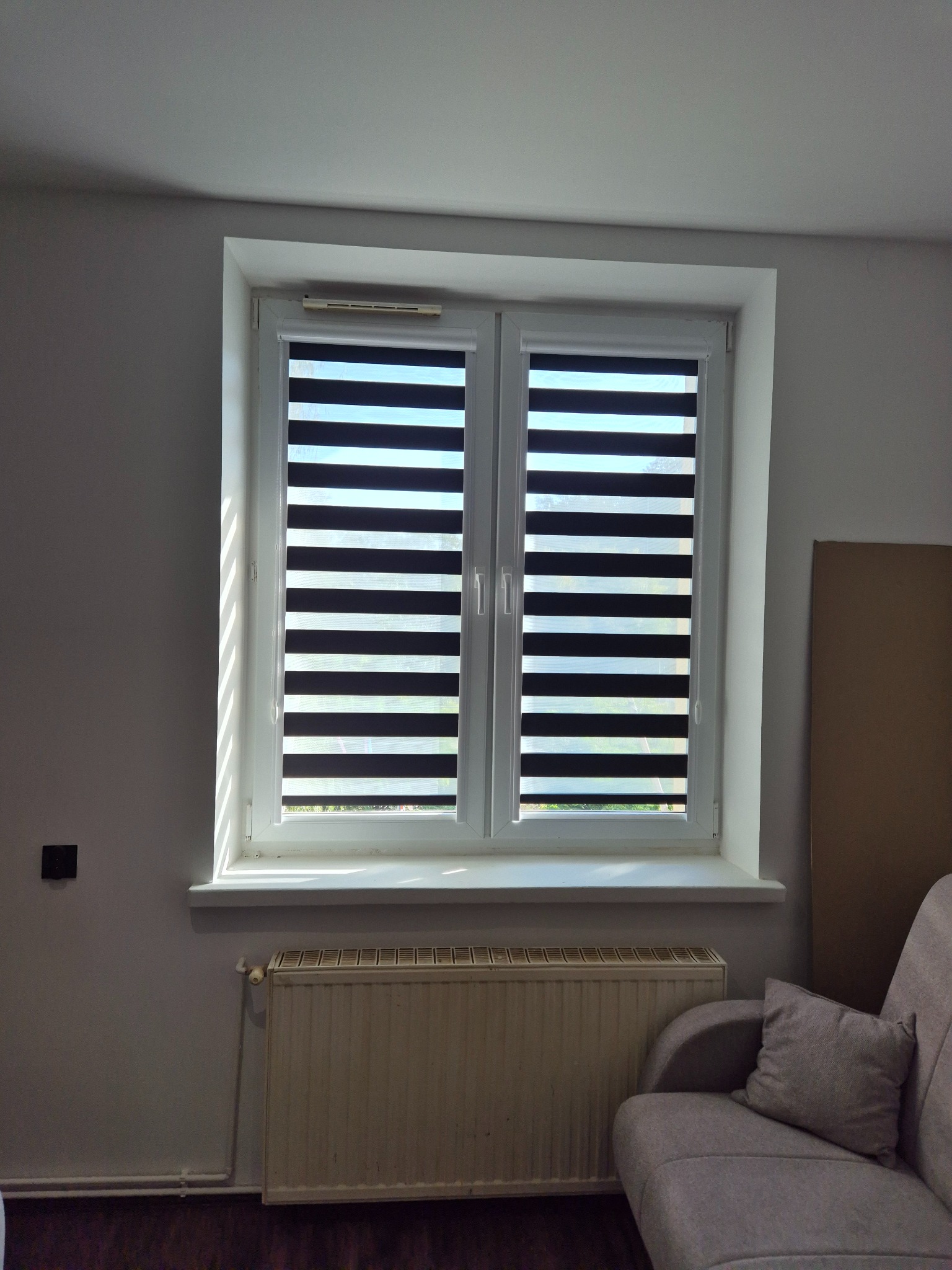 Okno z roletami dzień noc w białej ramie, widok z wnętrza. Na parapecie cień, pod oknem grzejnik, obok fragment sofy. Minimalistyczny wystrój.