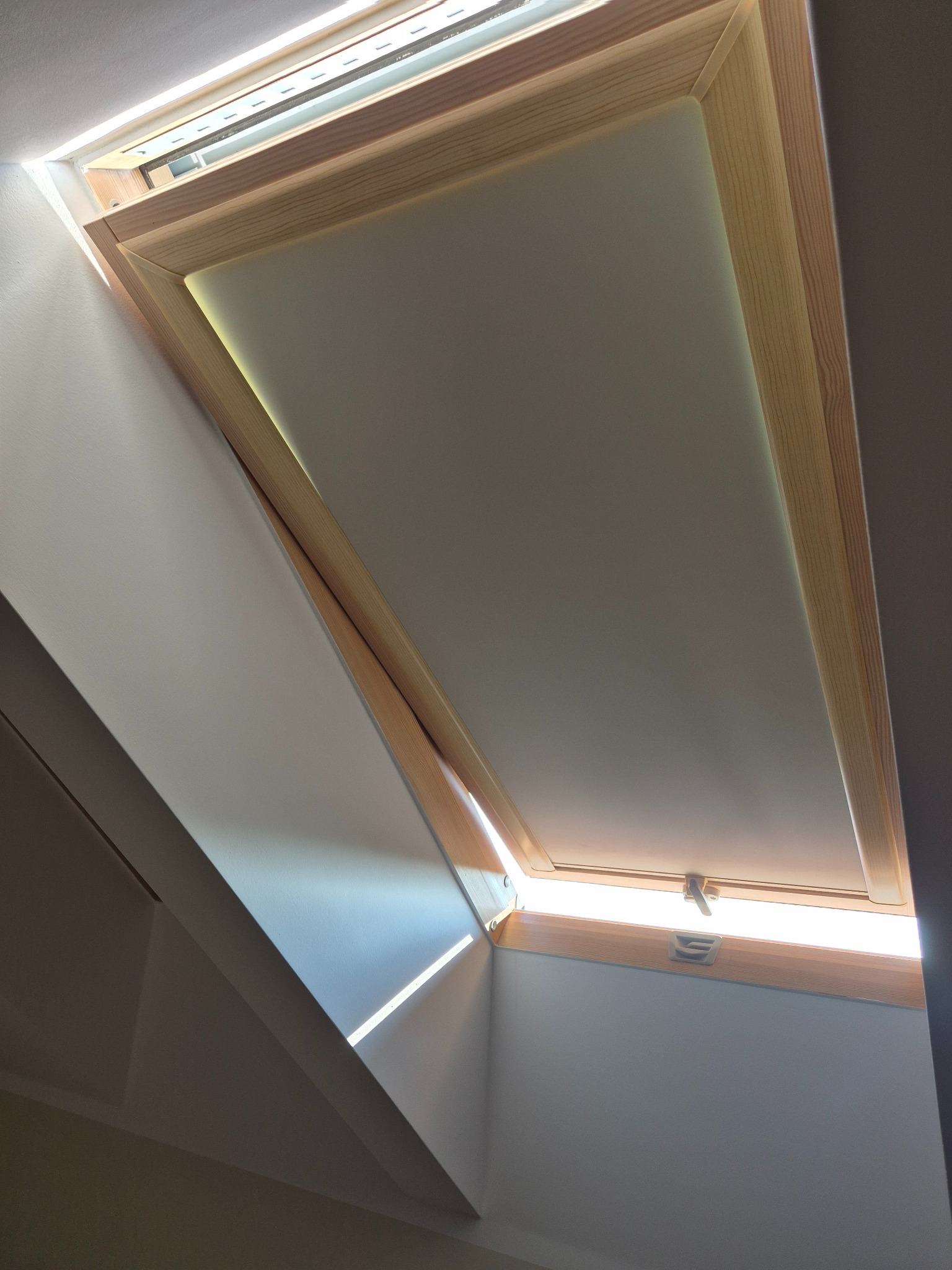 Drewniane okno dachowe z roletą w jasnym wnętrzu, uchylone, wpuszczające światło. Widok od dołu, ukazujący mechanizm i ramę okna.